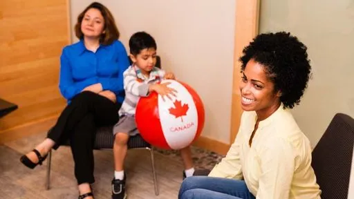 Two women sit in an office reception area. The woman on the left is accompanied by a young boy holding a beach ball styled like a Canadian flag.