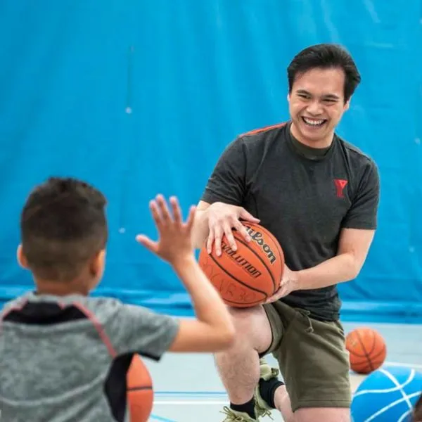 A volunteer YMCA basketball coach kneeling and holding a ball smiles at a youth player asking a question during a practice in a gynmasium