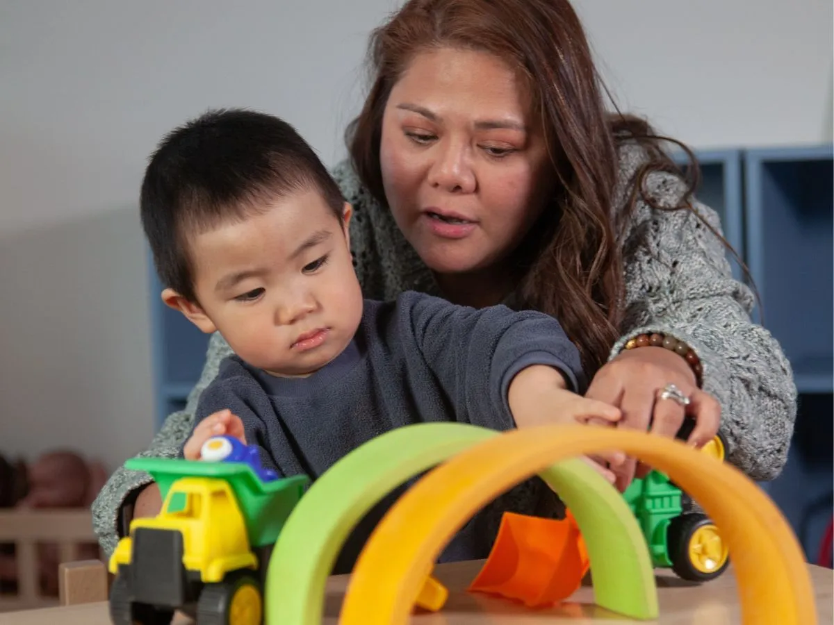 A woman helps a toddler play with a toy tractor on a table.